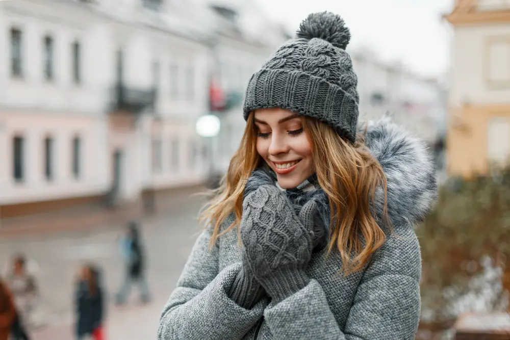 Happy attractive young woman in a winter knitted hat in a trendy gray coat with fur in knitted warm mittens with a beautiful smile