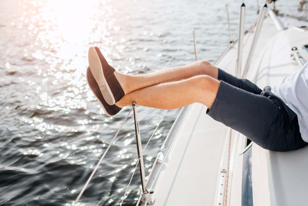 edge of yacht board. Guy wears dark shorts, white t-shirt and black boat shoes