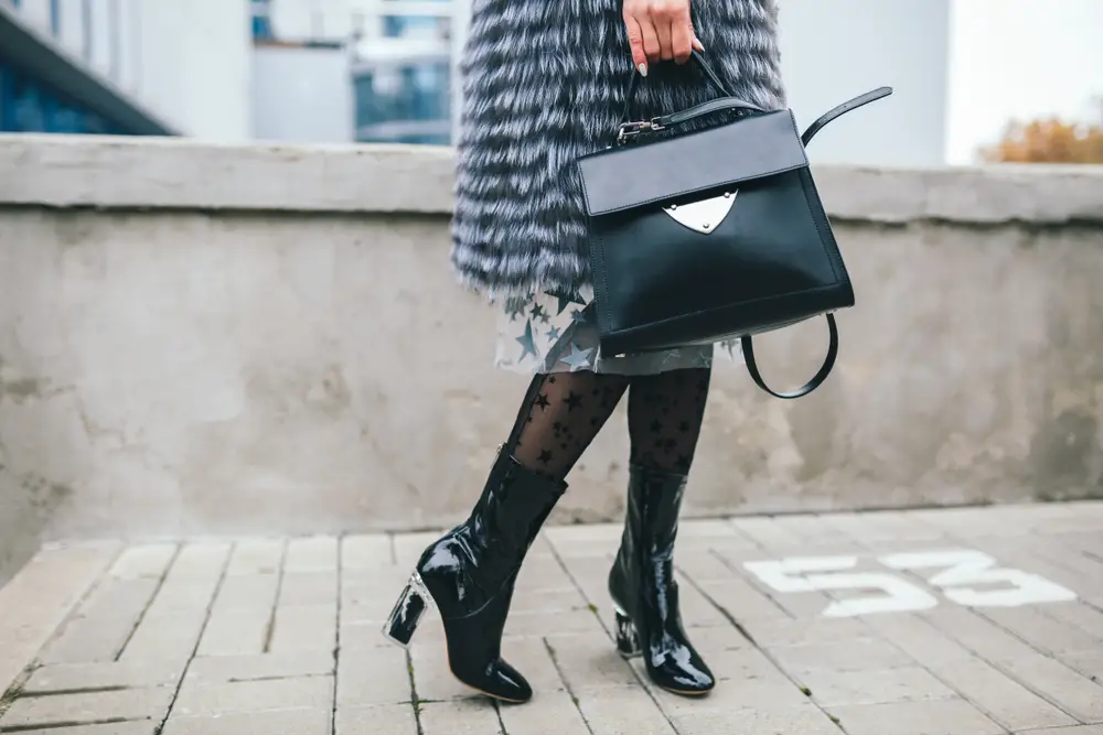 legs in calf boots of stylish woman walking in city in warm fur coat holding leather handbag