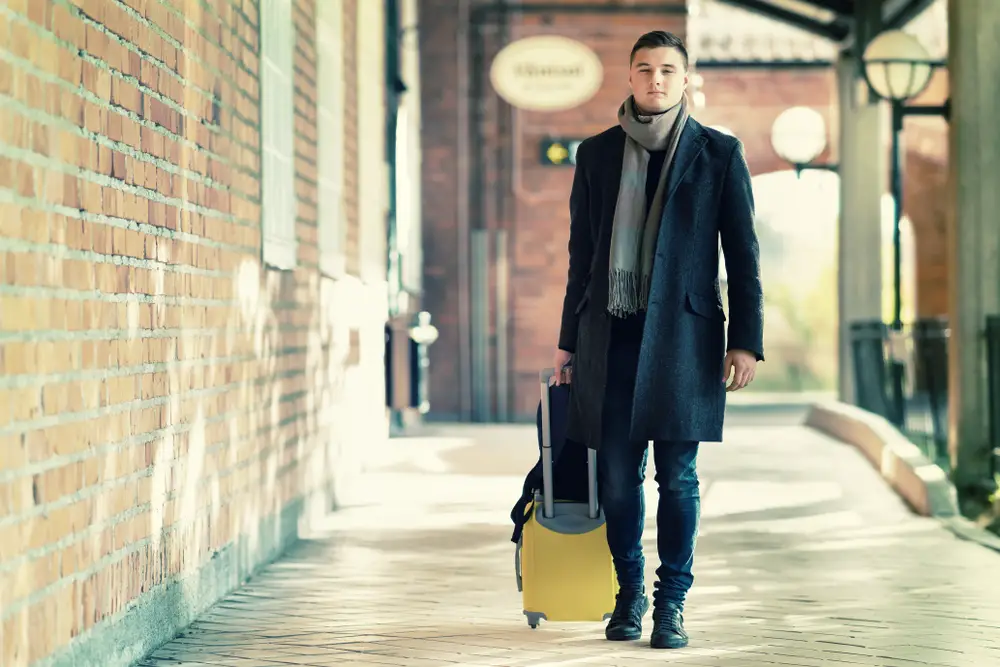 Young man in warm overcoat and scarf approaching a station with his suitcase in a concept of travel and vacations