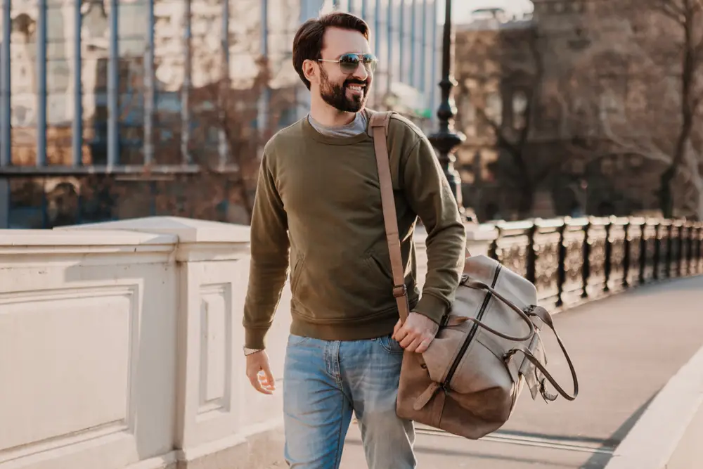 man walking in city street with weekender bag wearing sweatshot and sunglasses, urban style trend