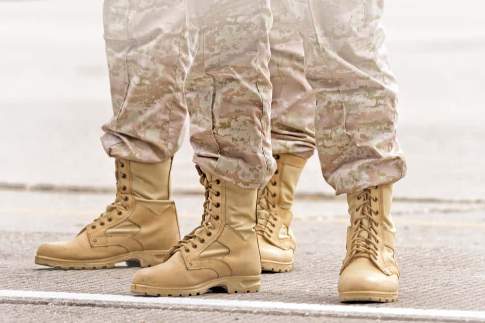 military boots on soldier feet army combat uniform on blurred background detail close up side view of leather shoe for hiking in sand color and brown camouflage pants