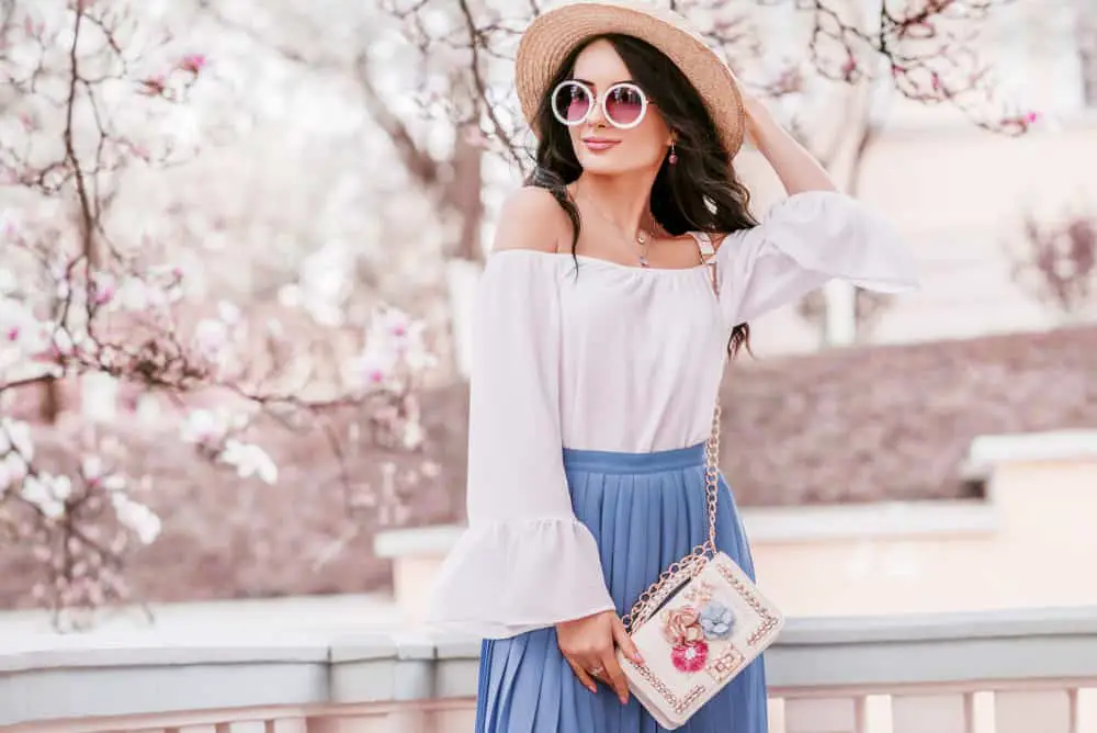 young beautiful woman wearing stylish round sunglasses, straw hat, cold shoulder blouse, pleated skirt, with small white bag